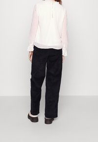 Světle růžová, texturovaná blůza s volány kolem límce a manžet, zapínání na knoflík na zadní straně; spárována s černými kordovými kalhotami a tmavými botami.