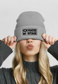 Gray knitted beanie with a prominent black rectangular patch featuring the text "ICH HASSE MONTAGE" and a cat graphic.