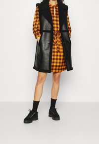 Černá kožená vesta s texturovaným límcem přes oranžové a černé kostkované šaty, stažená v pase. Stylizováno s černými ponožkami a robustními botami.