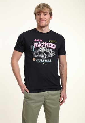 Młody mężczyzna o jasnobrązowych włosach, ubrany w czarną koszulkę z grafiką starego ciężarowego auta i kolorowym napisem "Rat Rod Culture".