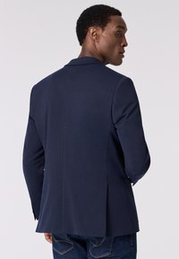 Blazer strutturato blu navy con tessuto testurizzato, spacco singolo sul retro e vestibilità sartoriale. Presenta rever classici e tasche frontali.