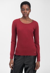 Maglione rosso a maniche lunghe con scollatura a girocollo, orlo e polsini a coste, realizzato in un materiale morbido, indossato con pantaloni neri strutturati.