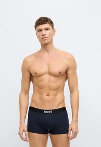 Granatowe bokserki z czarnym paskiem w pasie, na którym znajduje się napis "BOSS" w białym kolorze. Gładka tkanina i dopasowany krój. Model w pozycji stojącej.