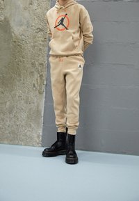 Felpa in pile beige con logo arancione e nero, abbinata a pantaloni jogging coordinati con un emblema. Indossata con stivali neri alla caviglia, suola texturizzata.