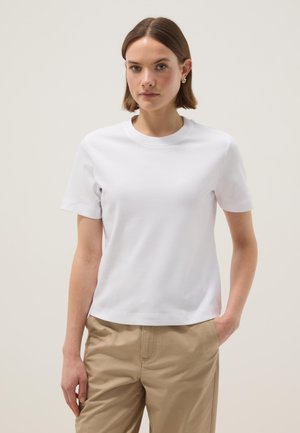 Jonge vrouw met kort bruin haar, gouden oorbellen, een eenvoudige witte T-shirt met korte mouwen en beige broek, met één hand in de zak.