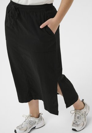 Fekete, oldalt sliccelt midi szoknyát viselő személy, amelynek zsebei vannak, fehér sportcipővel párosítva. Az egyik kezét zsebre teszi.