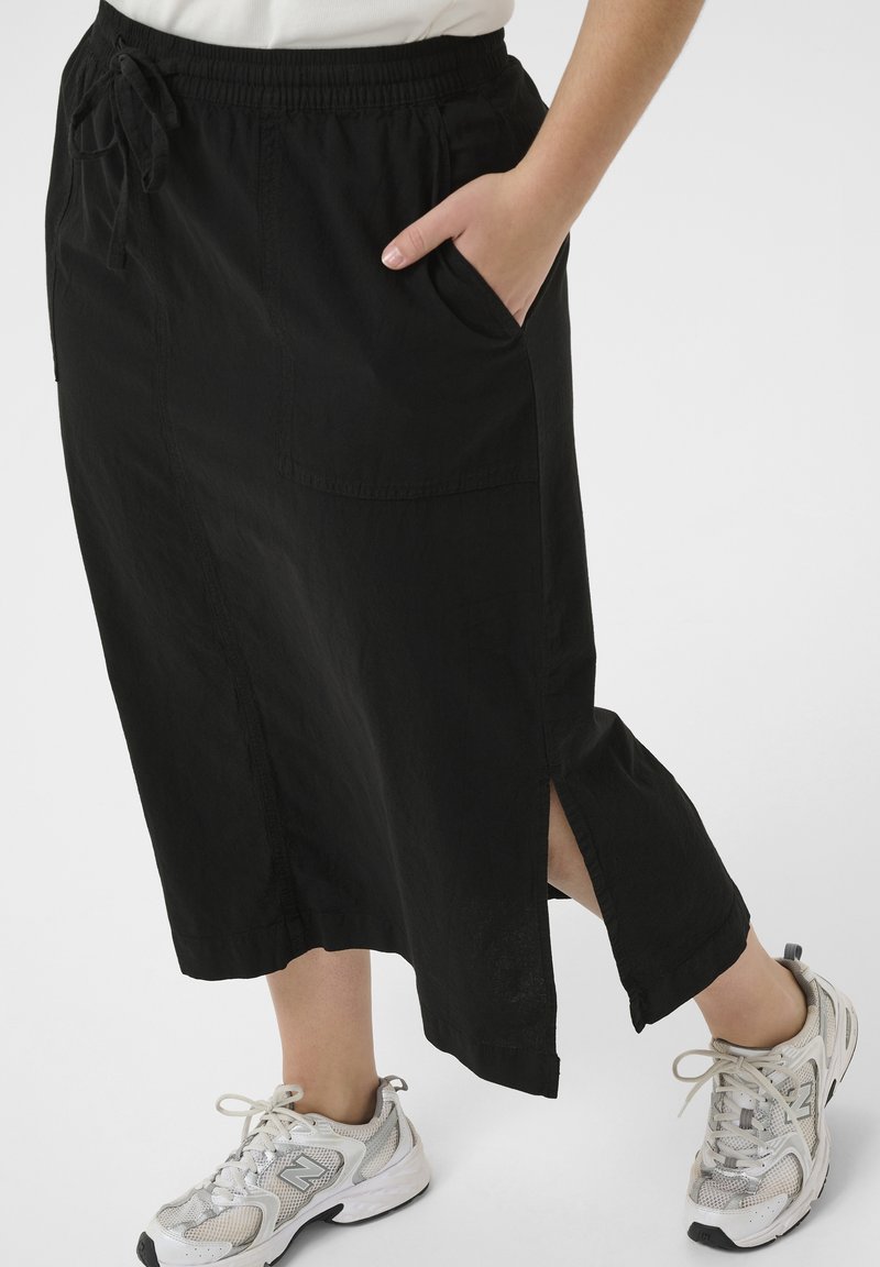 Personne portant une jupe midi noire avec une fente latérale et des poches, associée à des baskets blanches de sport. Une main repose dans la poche.