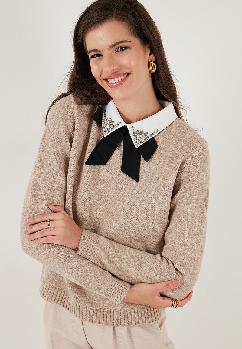 Giovane donna sorridente con un maglione beige con colletto bianco decorato e fiocco nero, pantaloni beige e orecchini e anelli d'oro.