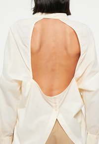 Chemise beige clair avec un dos ouvert, dotée d'une fermeture à boutons et de manches larges. Tissu en coton texturé, coupe décontractée.