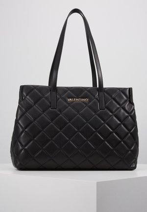Sac cabas Valentino en cuir matelassé noir avec double anse d'épaule, plaque logo dorée, présenté sur un socle blanc sur fond gris.