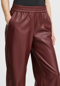Bordeaux faux leren broek met een elastische tailleband, voorzien van zijzakken en een gladde, glanzende textuur.
