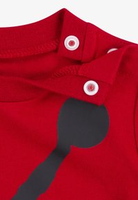 Camiseta de algodón roja con un diseño gráfico negro redondo. Cuenta con cierres de presión en el cuello con botones de plástico blancos. Textura suave.