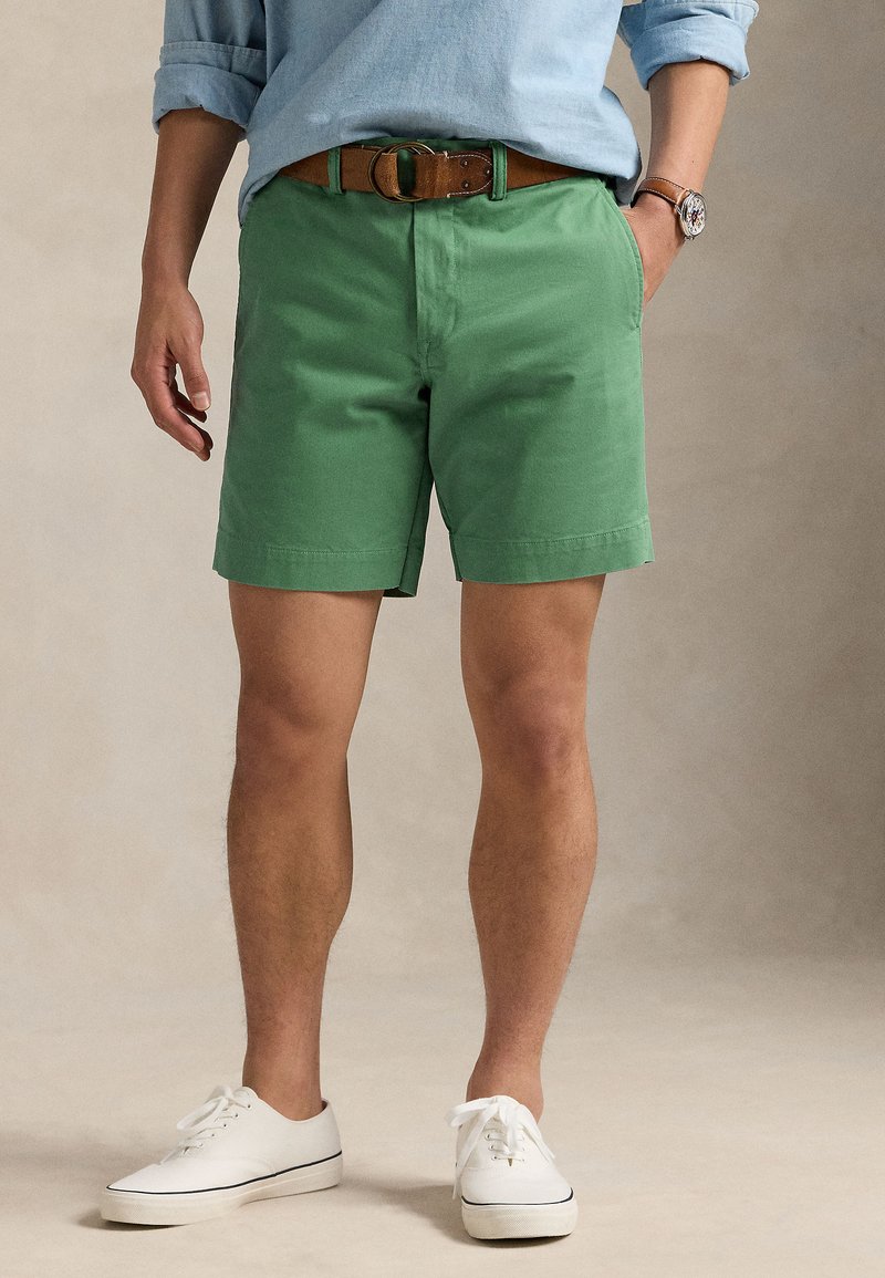 Zaļas kokvilnas šorti ar plakanu priekšpusi un sānu kabatām, kombinēti ar brūnu jostu un baltām sporta kurpēm. Vienkāršs dizains, ikdienišķa piegriezuma.