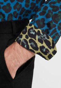 Een shirt met panterprint met een blauwe basis, zwarte vlekken en groene accenten; contrasterende gele manchetten met zwart panterpatroon.