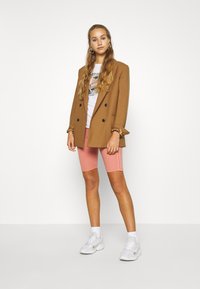 Blazer marrone doppiopetto sopra una maglietta grafica bianca, abbinato a pantaloni bike rosa e sneakers bianche. Capelli lunghi e mossi, grandi orecchini.