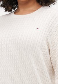 Maglione a maglia color beige chiaro con un motivo a treccia testurizzato, scollatura arrotondata e un piccolo logo in rosso, bianco e blu sul petto.