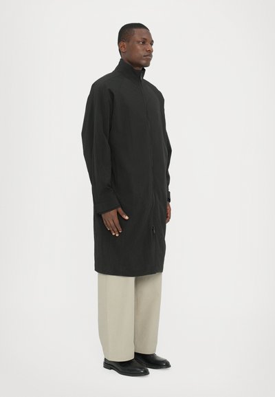 Black long-sleeve coat with a high collar and front zipper, made from a lightweight fabric. Worn over cream-colored wide trousers and black shoes.