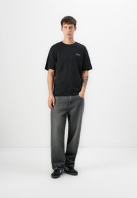 Svart bomull t-skjorte med korte ermer og en liten hvit logo på venstre bryst, sammen med grå, løse jeans og svarte joggesko.