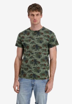 Mann trägt ein kurzärmeliges T-Shirt mit grünem Camouflage-Muster und hellblauen Jeans, steht vor einem einfachen weißen Hintergrund.