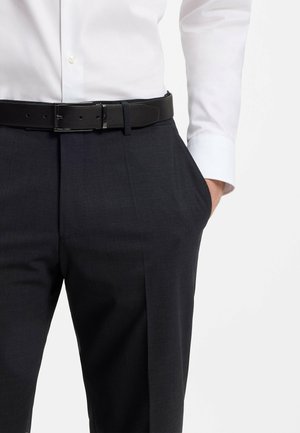 Hombre con pantalones gris oscuro, cinturón negro y camisa blanca de vestir, con la mano derecha en el bolsillo, sobre un fondo claro y liso.