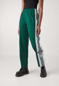 Vihreät verkkohousut, joissa on valkoiset sivuhiharesorit, elastinen vyötärö ja Adidas-logo. Valmistettu sileästä, kevyestä materiaalista, jossa on rento istuvuus.