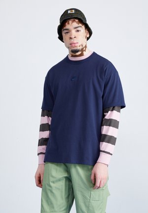 Camiseta de manga corta azul marino con un pequeño logo negro, superpuesta sobre una camisa de manga larga a rayas en rosa y marrón, combinada con pantalones cargo verdes.