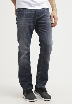Mand iført falmede blå jeans og sort-grå sneakers, stående med den ene hånd i lommen mod en ensfarvet hvid baggrund.