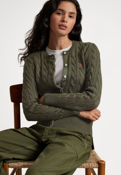 Πράσινη ζακέτα με πλεκτή πλέξη και στρογγυλή λαιμόκοψη, εφαρμοστά ριμπ μανίκια με μανσέτες και μικρή κόκκινη λεπτομέρεια λογοτύπου, συνδυασμένη με παντελόνι σε ελιάς πράσινο.