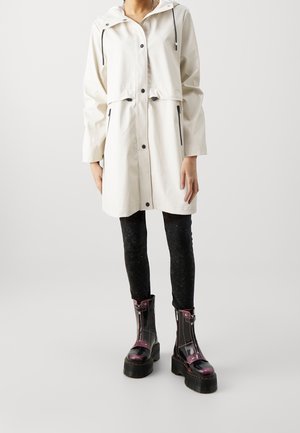 Manteau imperméable blanc avec capuche, lien de taille ajustable et fermetures éclair noires ; associé à des leggings texturés noirs et des bottines brillantes.