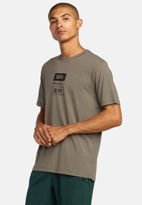Oliwkowa koszulka z krótkim rękawem w miękkim materiale, z czarnym chińskim napisem oraz logo RVCA nadrukowanym z przodu. Prosty dekolt typu crew neck.