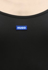 Κοντινό πλάνο του μαύρου λαιμού πουκάμισου με μικρό μπλε ορθογώνιο σημάδι με λευκό κείμενο "HUGO" στο κέντρο.