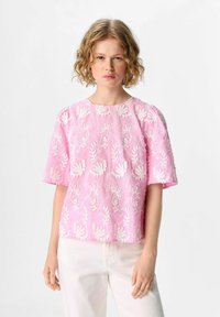 Haut rose brodé avec des motifs floraux blancs, à manches courtes et col rond, associé à un pantalon clair.