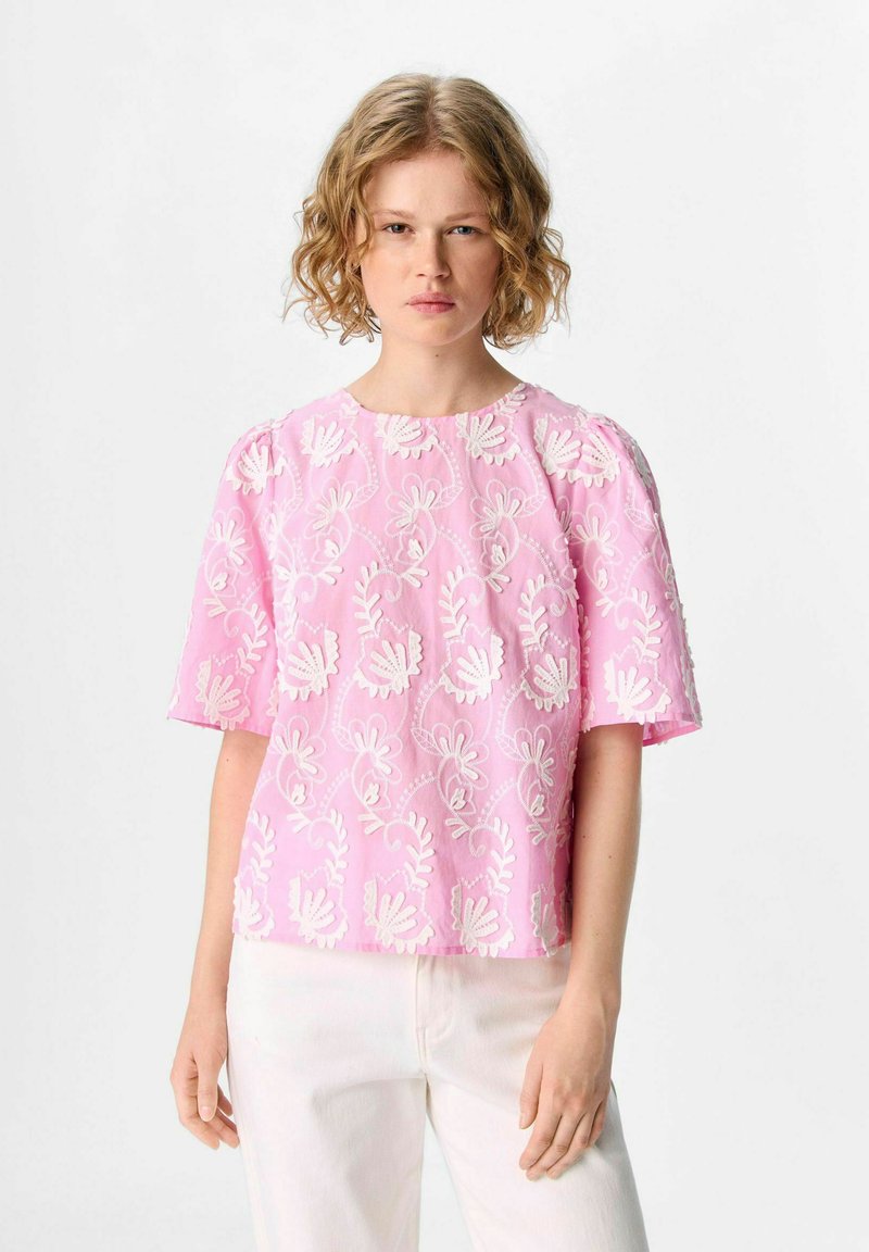 Haut rose brodé avec des motifs floraux blancs, à manches courtes et col rond, associé à un pantalon clair.
