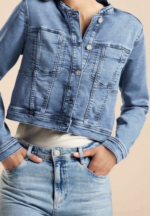 Mujer con chaqueta vaquera corta de color azul claro y vaqueros azules descoloridos de talle alto con las manos en los bolsillos, sobre un fondo neutro.