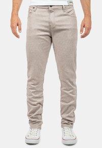 Beige Baumwollmischungshosen mit strukturiertem Gewebe, schlanker Passform, fünf Taschen-Design und schwarzem Knopfverschluss. Kombiniert mit weißen Turnschuhen.