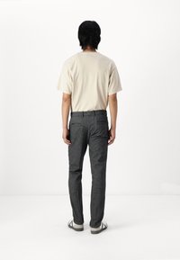 Beige Baumwoll-T-Shirt mit kurzen Ärmeln, kombiniert mit dunkelgrauen, strukturierten Hosen. Die Schuhe sind weiß mit grauen Akzenten. Rückansicht vor einem weißen Hintergrund.