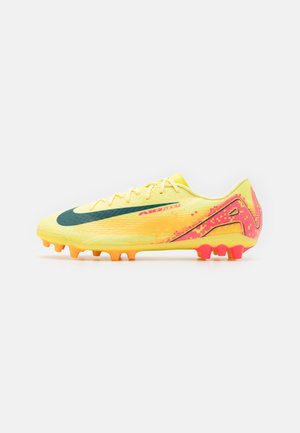 Botín de fútbol amarillo con un swoosh verde, acentos en rosa, una parte superior texturizada y una suela con tacos de punta naranja.