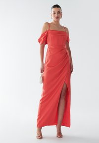 Lång korallfärgad klänning med axelbandslösa ärmar, strukturerad överdel, sidospår och draperat tyg. Accessoarer med en liten vit väska.