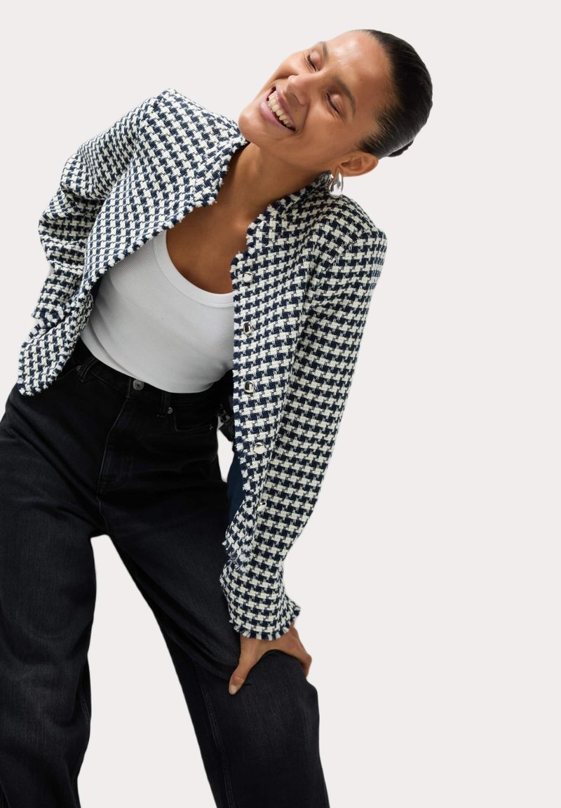 Blazer a scacchi navy e bianco con finitura testurizzata, dotato di colletto a volant e chiusura con bottoni, abbinato a jeans neri a vita alta.