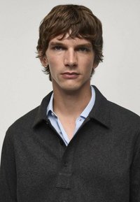 Giacca camicia grigio scuro realizzata in un tessuto testurizzato, con colletto, chiusura a bottoni e una camicia a righe blu chiaro sotto.