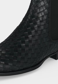 Bottine noire en cuir avec texture tissée et panneau latéral élastique, dotée d'un talon bas et d'un bout arrondi.