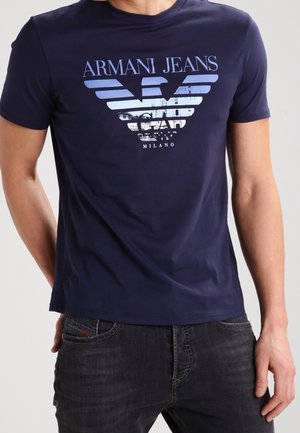 T-shirt Armani Jeans bleu marine avec grand logo bleu clair et blanc ainsi que le texte "Milano", porté avec un jean foncé par une personne debout.