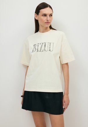 Kobieta o prostych ciemnych włosach ubrana w jasny, oversize'owy T-shirt z kwiatowym napisem i czarną mini spódniczką, stojąca na tle jednolitej białej ściany.