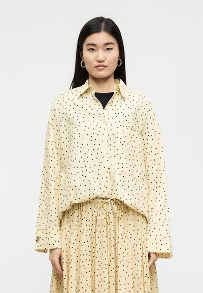 Γυναίκα με μακριά μαύρα μαλλιά φοράει μπεζ πουκάμισο με κουμπιά και πουά σχέδιο και φούστα που ταιριάζει με κορδόνι στη μέση, στέκεται μπροστά σε απλό φόντο.