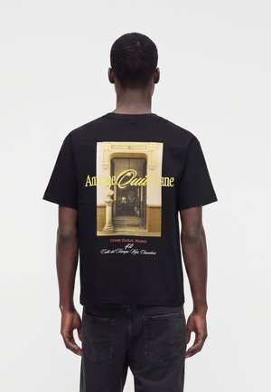 Homme portant un t-shirt noir avec une photo de porte architecturale vintage et un texte jaune et rouge dans le dos, debout devant un fond uni et clair.