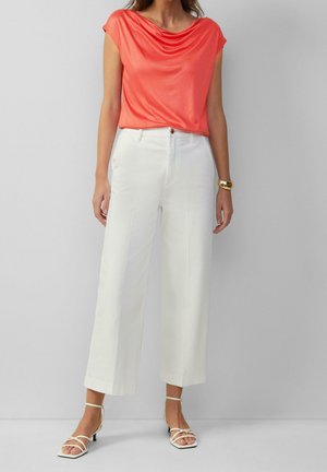 Vrouw draagt een koraalroze gedrapeerde blouse met korte mouwen, een wit wijduitlopend broek met hoge taille, witte sandalen met bandjes en een gouden armbanden cuff.