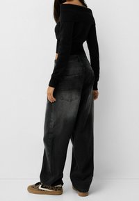 Pull noir à épaules dénudées associé à un jean en denim gris foncé taille haute à jambes larges avec un léger effet délavé, complété par des baskets à motifs.