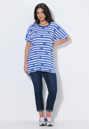 Vrouw staat met hand op heup, draagt een oversized blauw-wit gestreept shirt, donkere opgerolde jeans en witte sneakers met roze details.