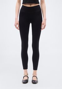 Μαύρο κολάν με ψηλή μέση, κατασκευασμένο από λείο, ελαστικό ύφασμα. Φορεμένο με μαύρα επίπεδα παπούτσια, παρουσιάζοντας ένα κομψό και εφαρμοστό σχέδιο.
