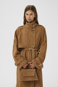 Cappotto trench in suede marrone con colletto alto, chiusura doppiopetto, cintura e ampie maniche, abbinato a una piccola borsa coordinata.